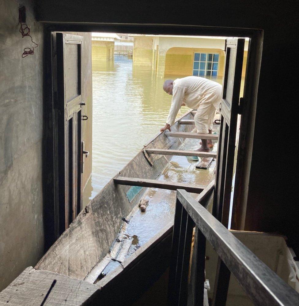 Mohammed Sani Gambo stands in a canoe. Its front is moored inside the doorway of his flooded house.