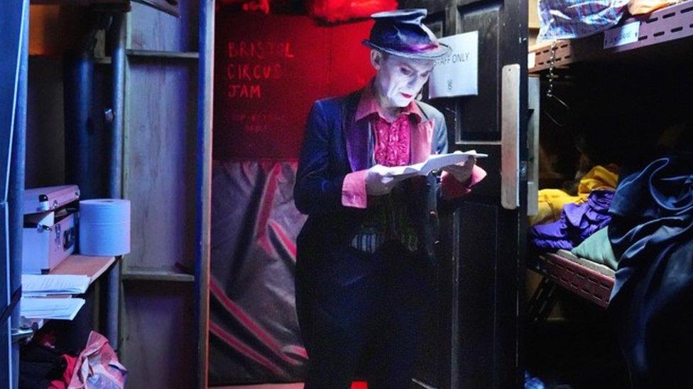 Doug Francis in costume, make up and a flattened hat in a dark, small cupboard room looking at a piece of paper