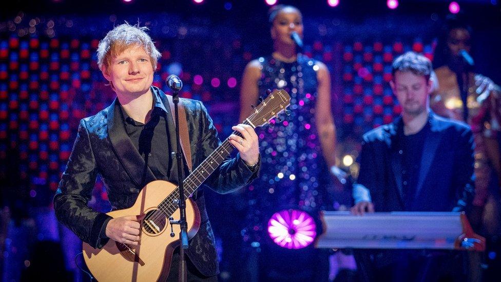 ed sheeran holding a guitar with a pianist playing in the background