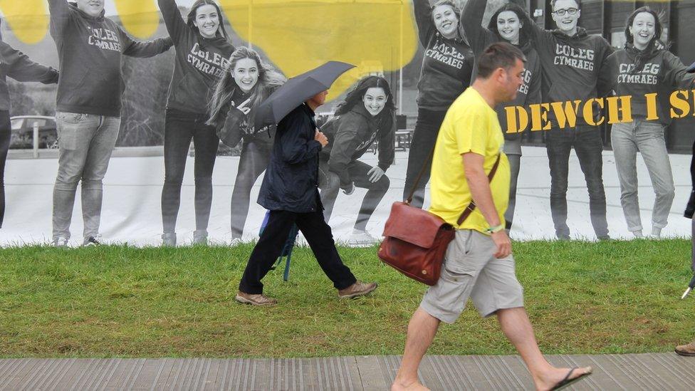 Dau ddyn yn cerdded i'r run cyfeiriad, un mewn trowsys byr a sandalau, a'r llall efo ambarèl // Two men walking in tandem - one in shorts and flip-flops, the other in full length trousers and umberella