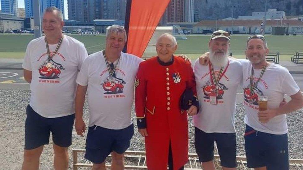 Four men wearing white t-shirts and medals stand either side of a Chelsea pensioner.