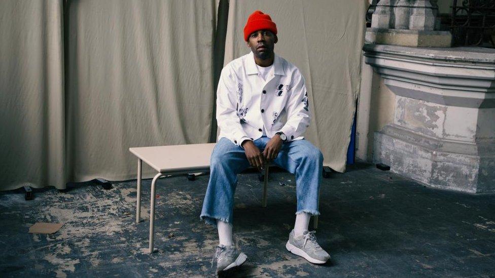 House of EL with short dark hair wearing a red hat and white shirt sitting on a table