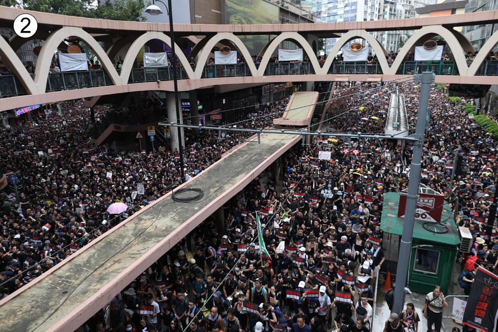 The street can no longer be seen beneath the heaving crowd in this shot of a light rail station, the ring and arches of its architecture visible above them