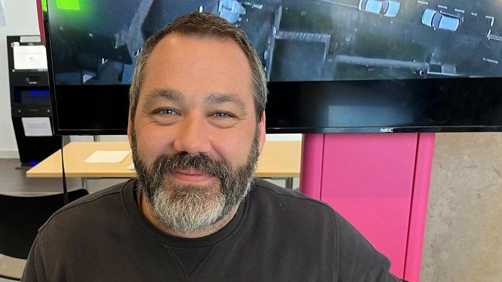 Man with short hair and beard sits in front of a TV screen