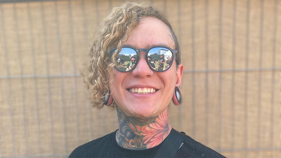 Will Ramos smiling for the camera. He is wearing sunglasses and has his curly blonde hair to the one side of his head. He has bright neck tattoos and is wearing a black T-short