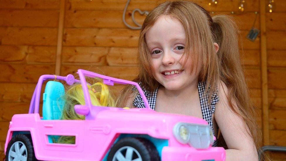 A little girl sits at a table with a toy car. She is smiling at the camera.