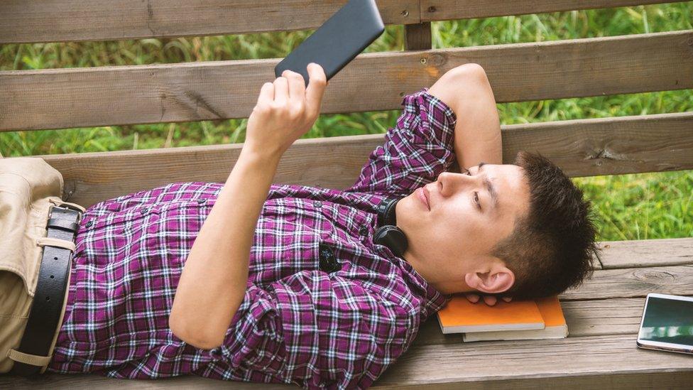 Man reading a digital tablet lying on bench with books under his head