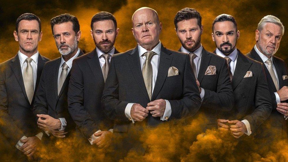 Six major EastEnders male characters stand in a line with stern expressions surrounded by golden smoke. From the left, there's Jack Branning, Nish Panesar, Dean Wicks, Phil Mitchell, Keanu Taylor, Ravi Gulati, and Rocky Cotton. Each man wears an identical grey/black suit with white shirt, gold necktie/pocket square combo and gold cufflinks.