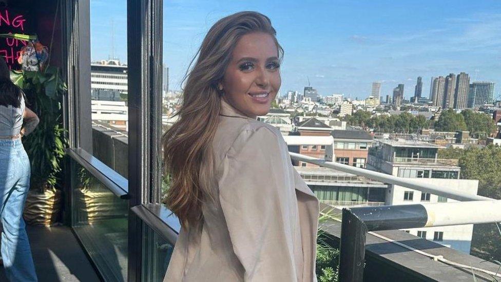 Molly Plumb posing for a photo near a window with a view of a skyline. Her body is facing out to the window but she is turning her head over her right shoulder to look at the camera and smiling. She is a white woman with make-up on and has long blonde hair. She is wearing a long-sleeved beige shirt or coat.