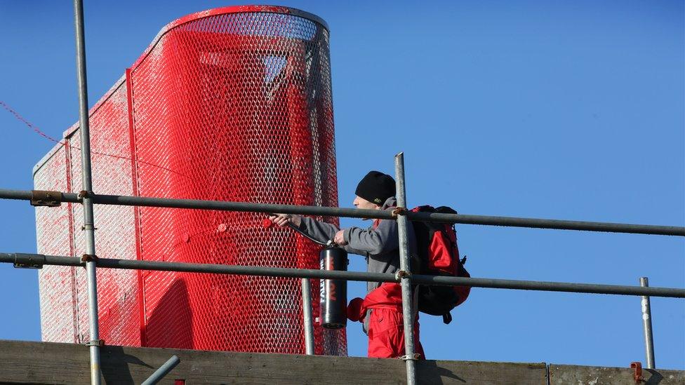 A man on a rooftop sprays red paint over a large metal structure