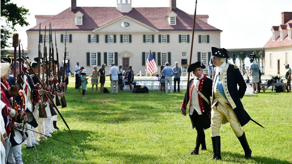 People in historical dress are pictured with bayonette weapons on lawn in front of historical estate