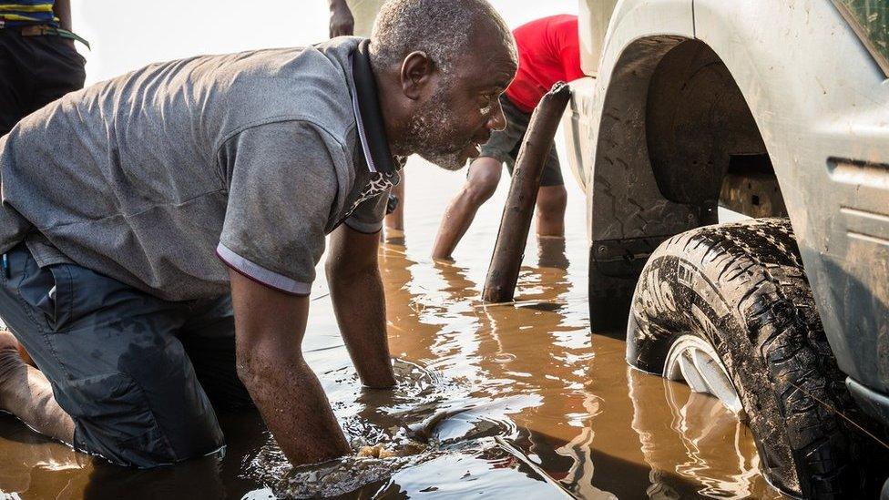 Man knee-deep in water looks under the wheel arch of a stranded car