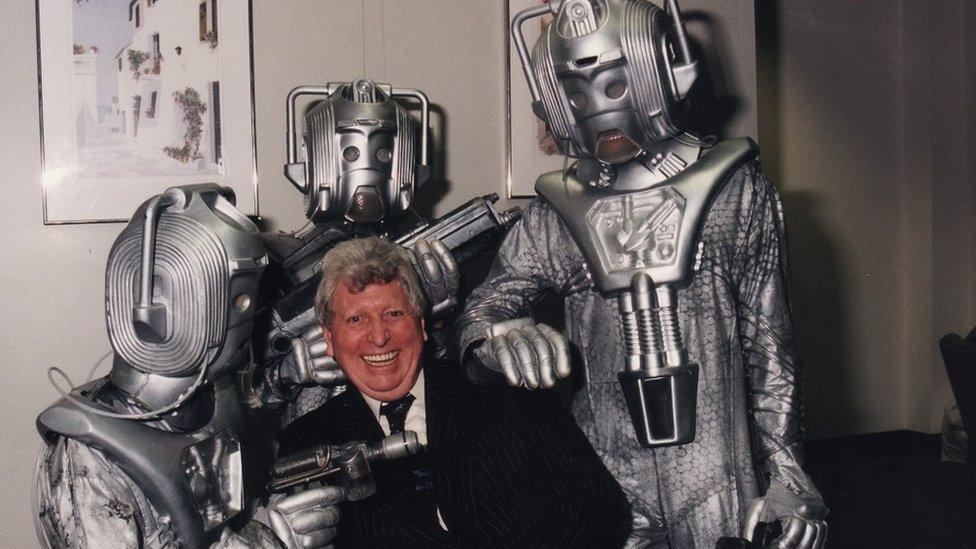 Tom Baker siling at the camera, surrounded by Cybermen