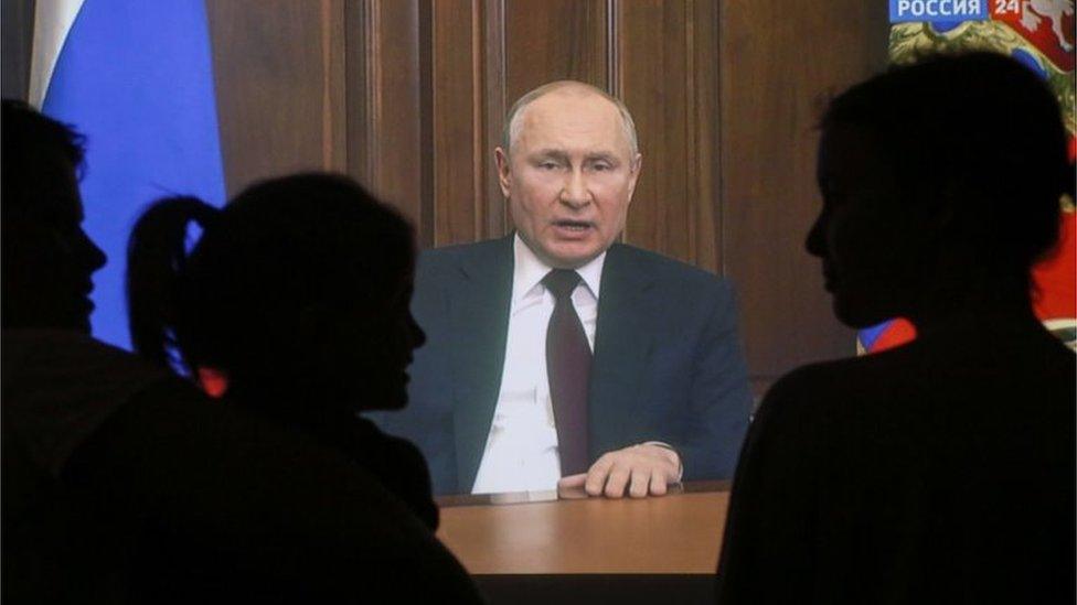 A family are silhouetted against a massive screen playing Putin's address