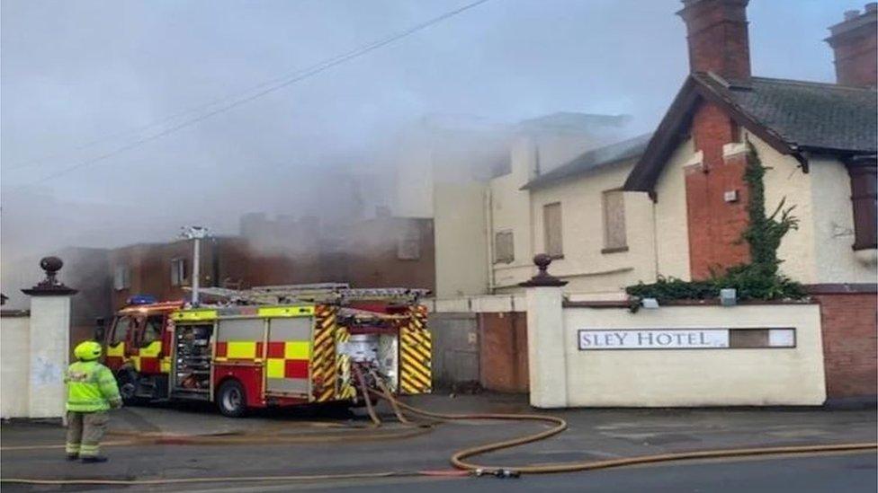 Fire engine outside a building with boarded up windows and plumes of smoke visible