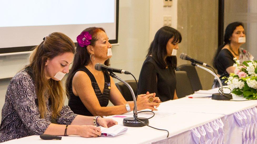 Four women with their mouths taped shut in front of microphones