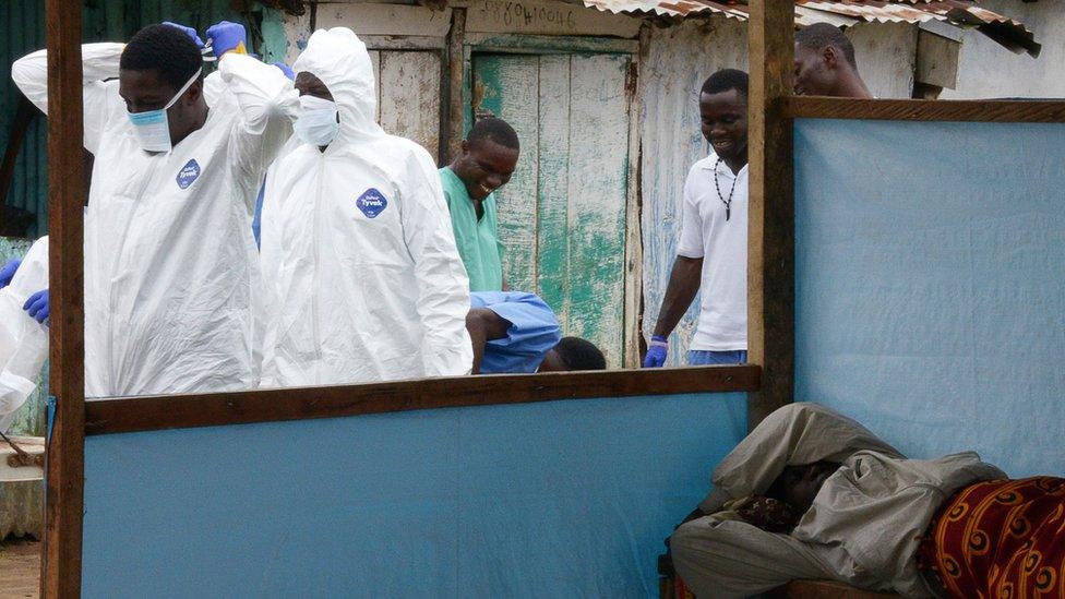 Ebola medical workers in protective suits walk past a sick women