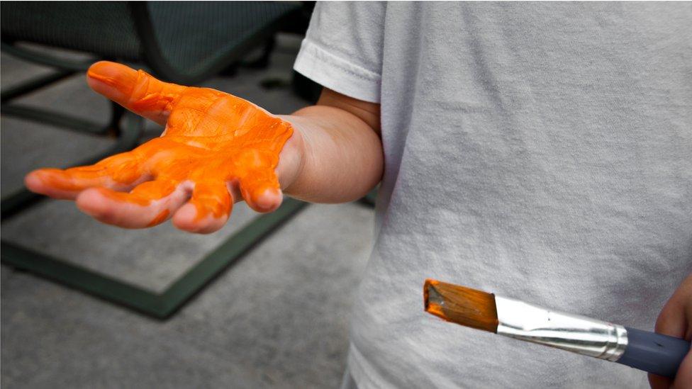 Boy with orange paint on one hand and holding a paint brush with another