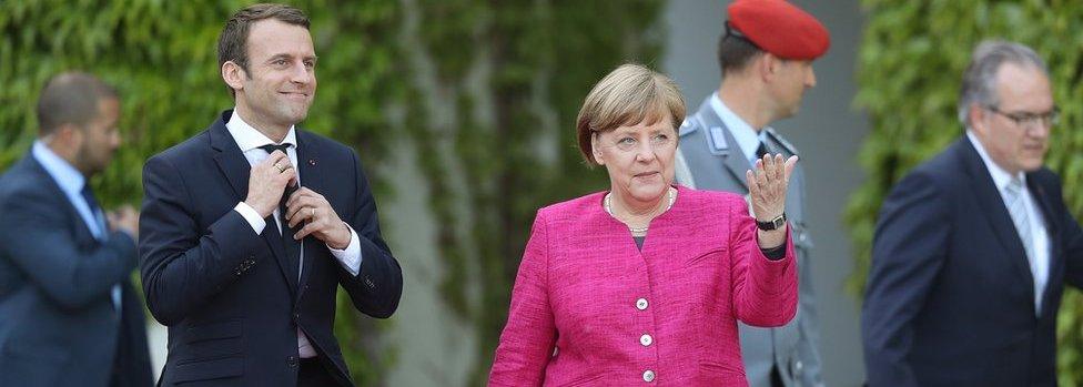 Emmanuel Macron and German Chancellor Angela Merkel walk to review a guard of honour upon Macron's arrival at the Chancellery (15 May)