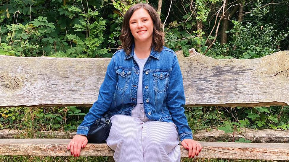 Sophie, sitting on a wooden bench, wearing a light coloured top and trousers and a blue denim jacket. behind her is greenery.