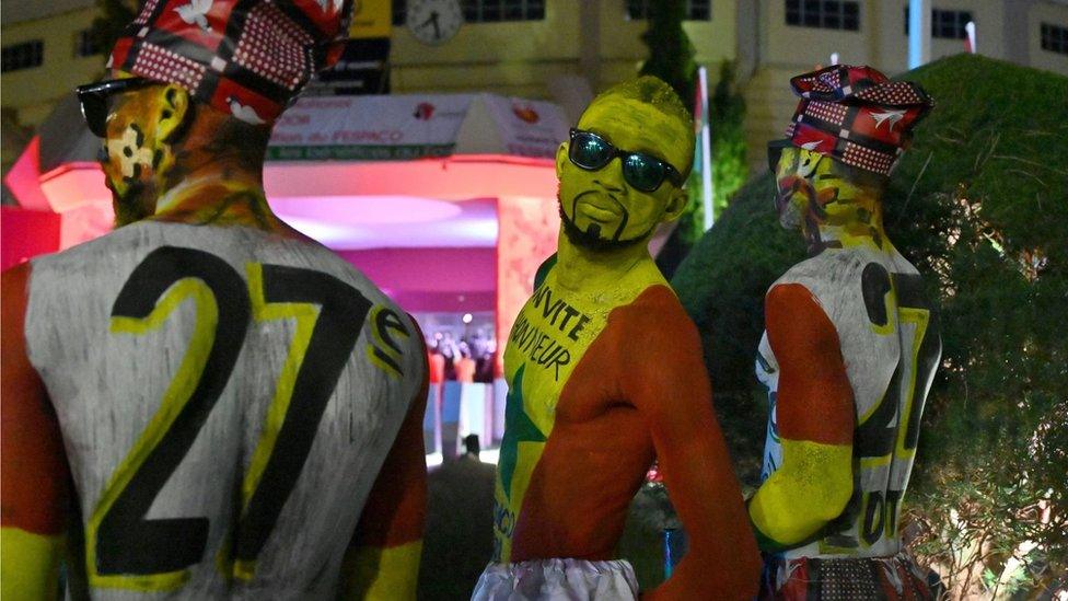 Men standing with colourful body and face paint. They have the colours of the Burkina Faso flag