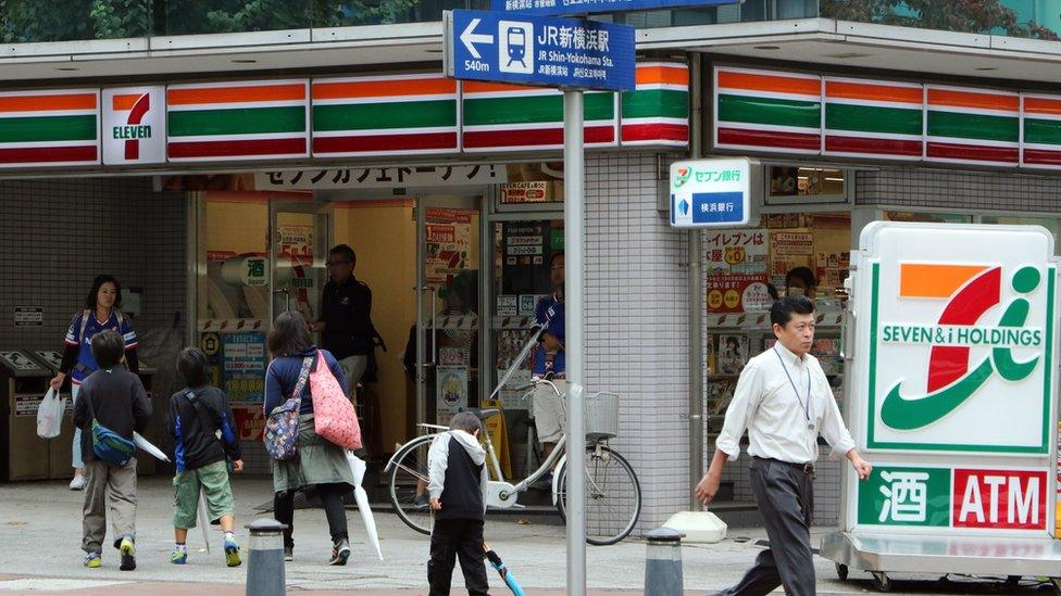 Exterior of a Japanese 7-Eleven, with three adults and three children walking in front of it