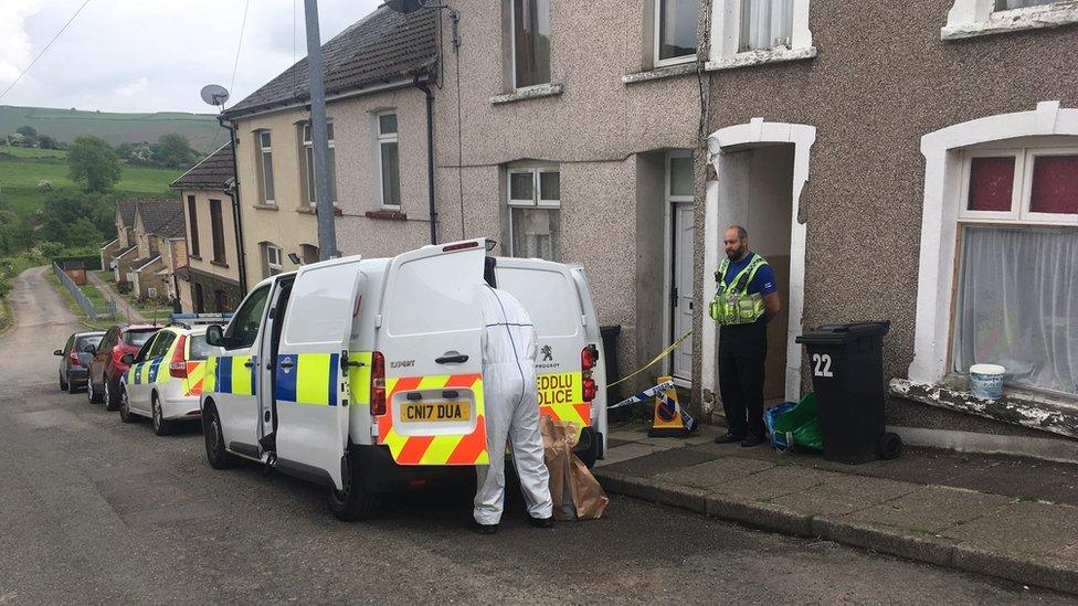 A row of terraced houses - an officers stands guard in front of a house where there is a wheeley bin with the number 22 on it. A forensic officer in forensic clothing is looking in the back of a police van