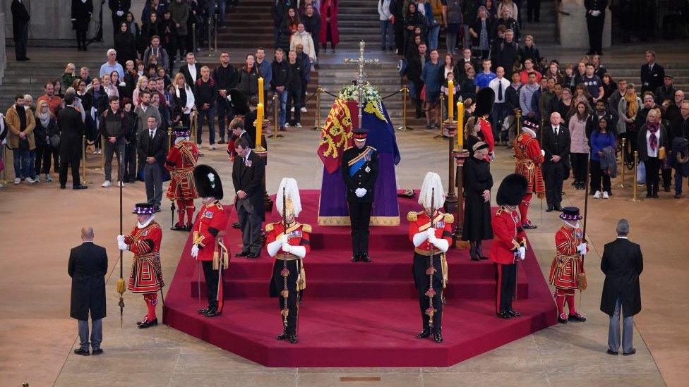 The Queen's coffin, surrounded by her grandchildren watching guard, as well as royal guards and members of the public behind.