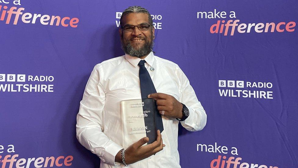 A man in a shirt and tie standing in front of a purple branded banner holding an award made from glass.