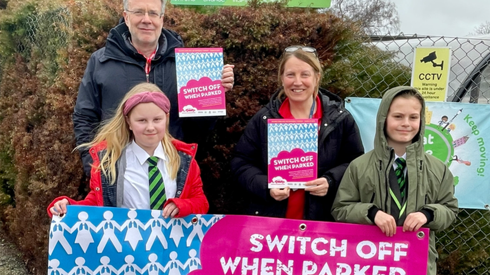 Paul West and two pupils holding a "switch off when parked" banner