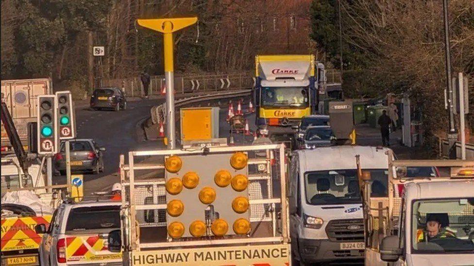 Roadwork vans and trucks around the centre of a road with a silver pole standing in the centre and yello T-shaped camera on its top.