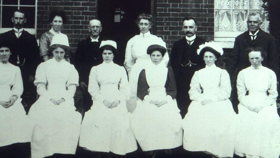 A black and white image of the staff of Thetford Workhouse in the about the early 1900s. There is a row of six white-clad women sitting down and a row of four men and two women are standing behind them. They are wearing Edwardian dress