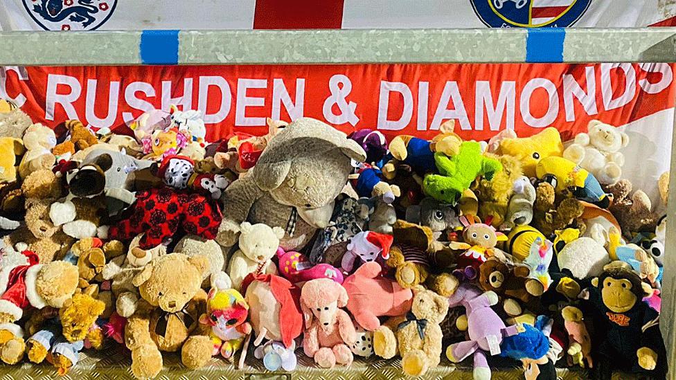 A mound of teddy bears and other soft toys, backed by an England flag with Rushden & Diamonds in white capitals across the horizontal red stripe.