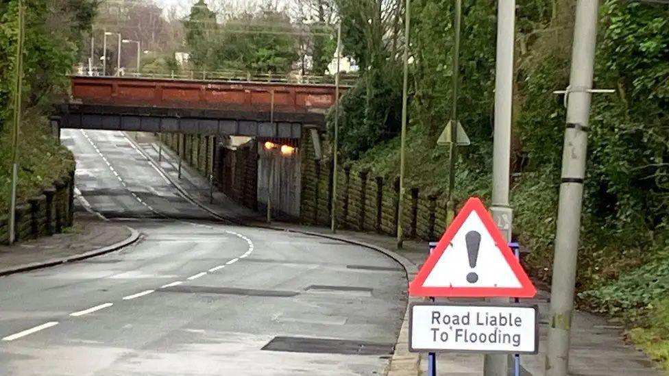 A road goes down into a dip under a railway bridge. A triangle shaped warning road sign with an exclamation mark and a sign below says road liable to flooding.