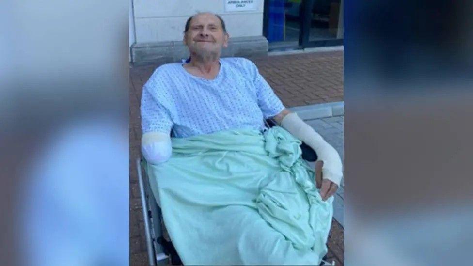 Vic Franklin smiling in a wheel chair while wearing a light blue blanket and a hospital gown. He is a elderly white man who is bald on top with dark hair on the sides of his head. He has a bandage on his right arm which has been amputated by the elbow and a bandage on his left arm and hand.