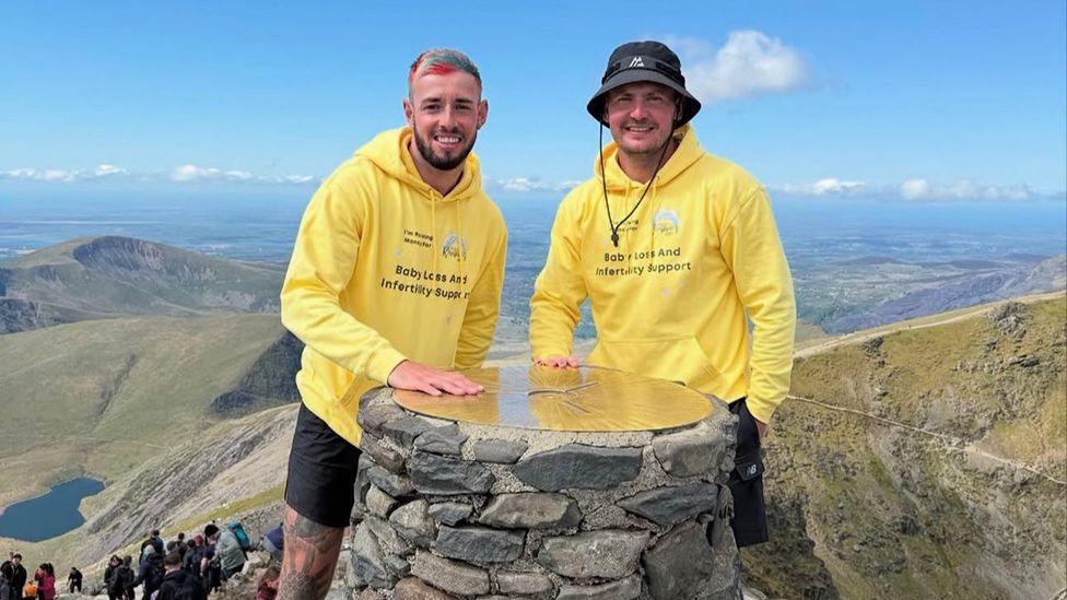 Joel Redhead with multi-colour hair and a black beard stands next to his friend Shane who is wearing a blue hat on the summit of Snowdon. The pair are wearing yellow hooded tops with the Chasing Rainbows charity logo on them.