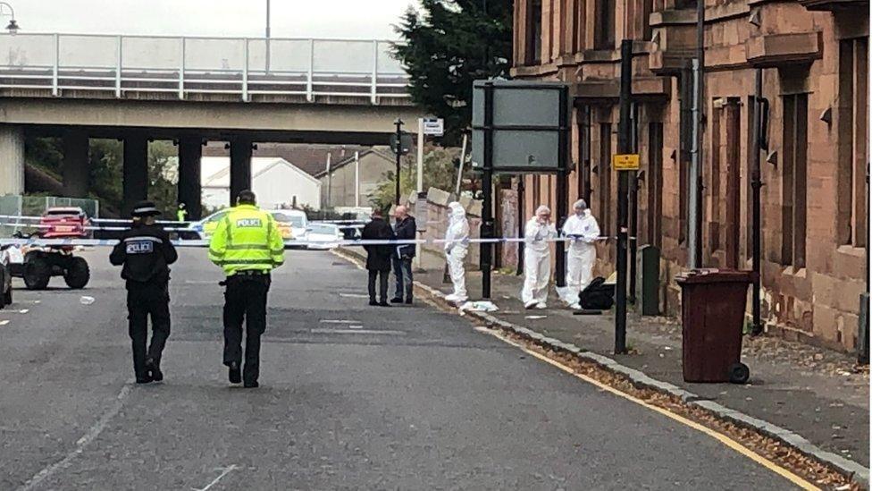 Police in hi vis clothing an forensics officers in white overalls walk around police tape outside a red sandstone tenement building.