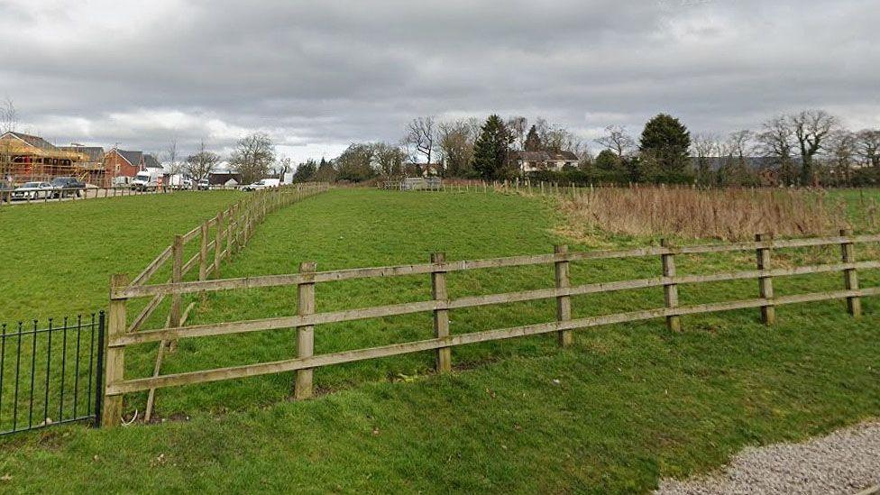 A large fenced field with new red brick houses being built in the distance. The horizon is tree-lined on an overcast day.