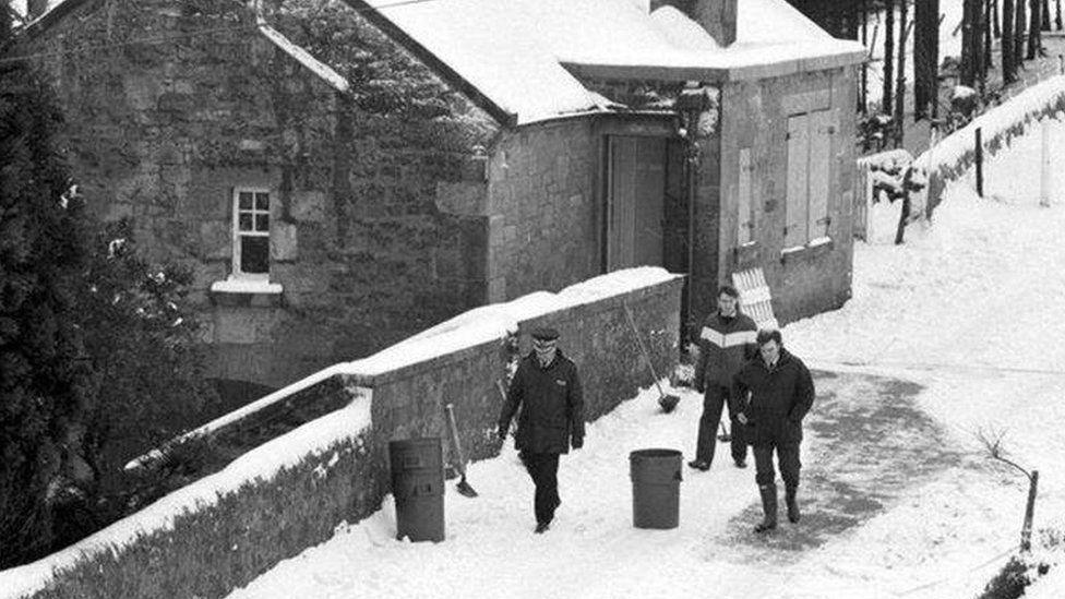 This black and white photograph shows a policeman and two other men walking in the snow past Loganlea Cottage. There is thick snow on the ground and along the wall. There are spades resting against the wall and two large buckets.