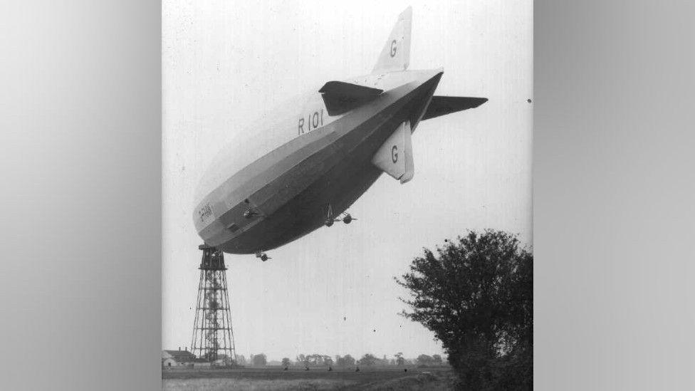 A black and white image of a huge airship which is tethered to a large tower. It is floating above a field with grass and trees in it. On the side of the airship it says "R101" and it has small wheels on the bottom of it