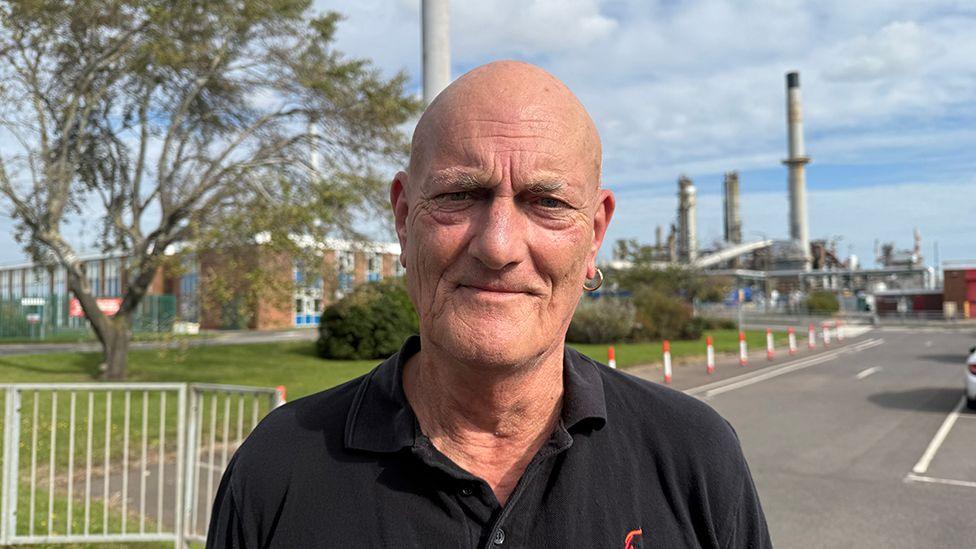 Mick Simpson from the union he has a shaved head and an earing in his left ear he is wearing a dark blue polo shirt with the Unite logo on it and is standing outside the refinery