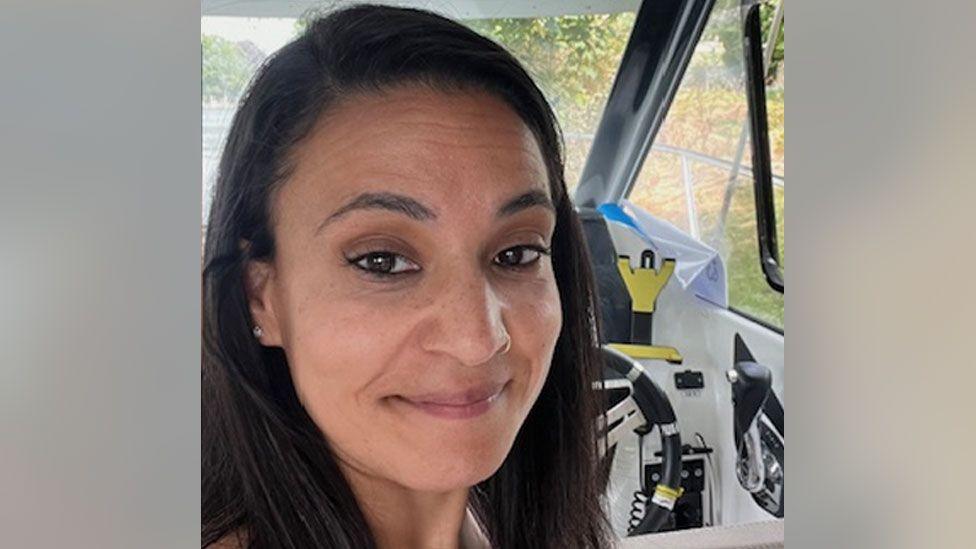 Syreeta Sandhu smiles at the camera. She is in the driver's seat of a car