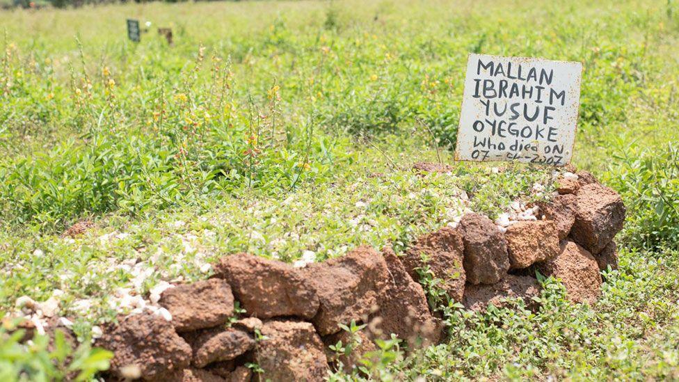 A grave - a raised bed - surrounded by stones and green vegetation in Tudun Wada cemetery. At the top of the grave is white sign with a hand-written inscription naming the person who died