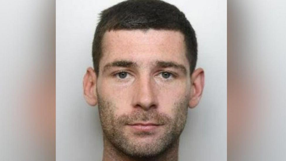 Michael Trainer's mugshot, where he is staring at the camera with a serious expression and has dark brown hair and dark stubble. He is stood in front of a light grey background.