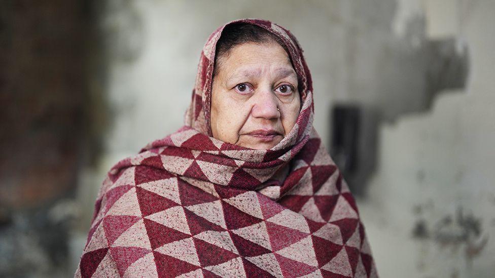 Bushra Rashid, wrapped in a thick shawl with a red and beige geometric triangle pattern, is standing against a worn, textured wall with patches of peeling paint and discoloration.