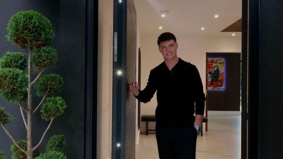 Billy stood in his doorway, with a plant on the left side and artwork out of focus in the background.