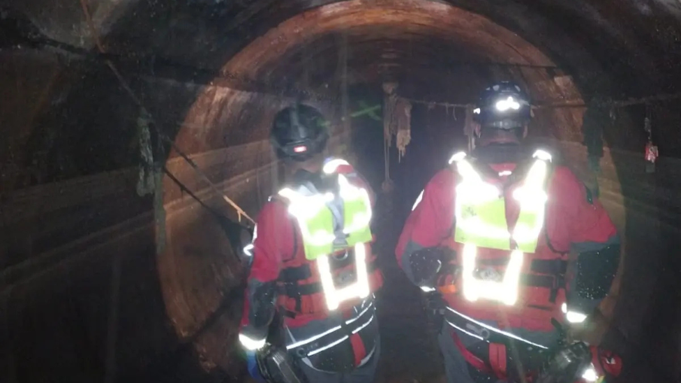 Two members in high vis jackets, and helmets walking in an underground sewer.