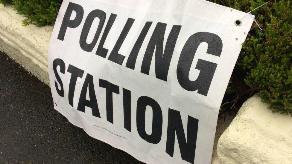 A canvas sign saying "Polling Station" attached to a low wall and a hedge.