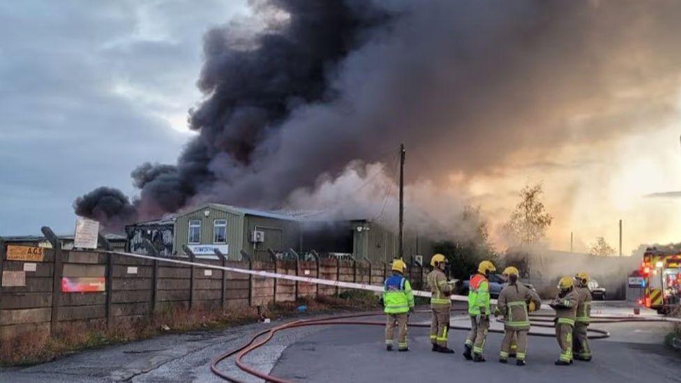 A group of firefighters in brown uniforms with yellow helmets stand near a concrete fence at the side of an industrial estate, where a large plume of black and grey smoke rises from green buildings