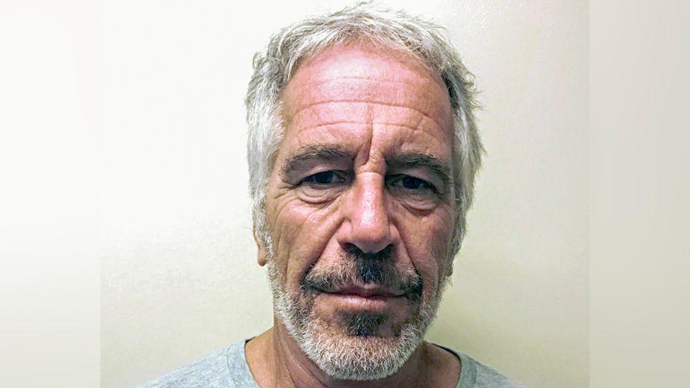Close-up portrait of Jeffrey Epstein taken in jail in 2019, with short grey hair and a grey beard, facing the camera against a plain light-coloured background. The image is tightly cropped around the head and shoulders, with even indoor lighting and no visible text or other objects.